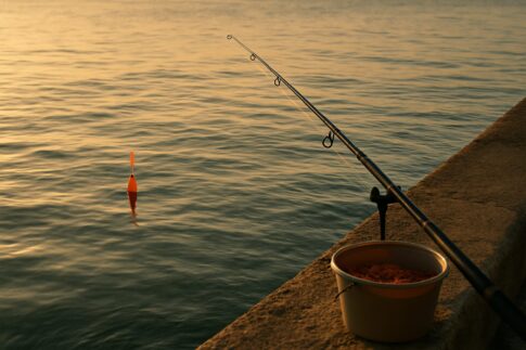 堤防のそばで水面に浮かぶオレンジ色の円錐ウキと磯竿のフカセ釣りシーン